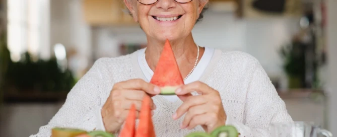 Smiling older adult holds a slice of watermelon at a table filled with fresh fruit, showing senior nutrition tips at The Village Senior Living.