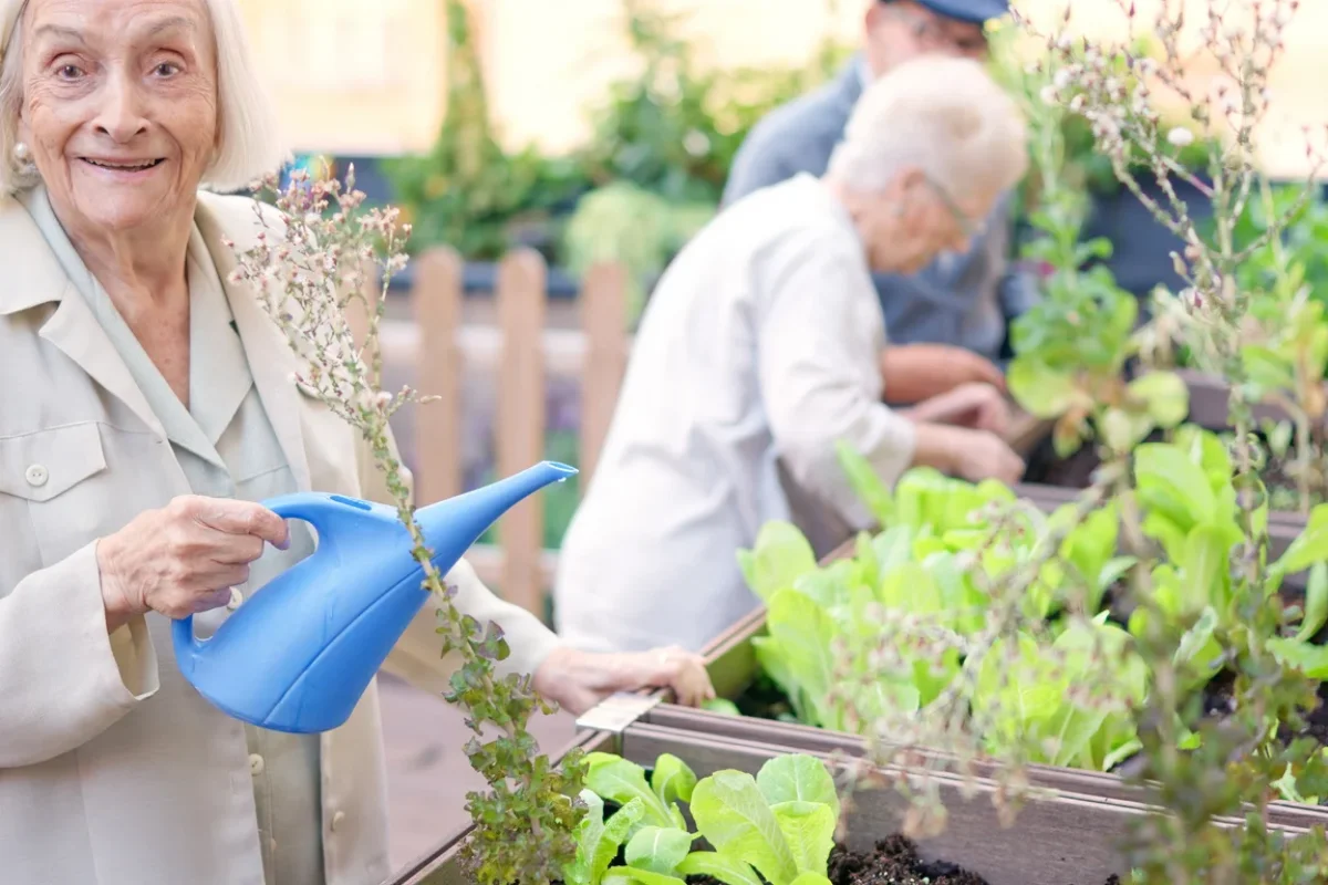Memory Care vs Assisted Living Senior Garden Support Older woman smiling while watering plants in a garden, representing memory care vs assisted living and supportive senior living options.